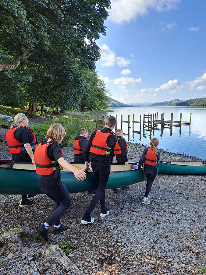 Image description: Six young people carry a canoe down a rocky shore to a lake